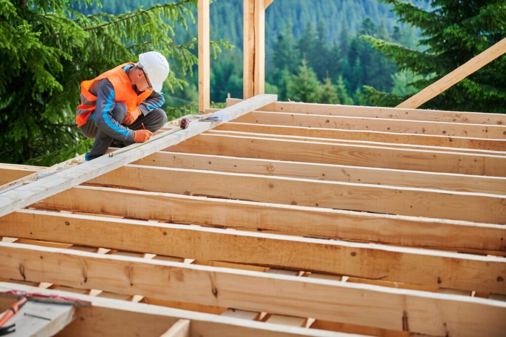 Carpenter in the process of building wooden frame house. Man measures distances with a measuring tape while dressed in workwear and helmet. Concept of modern, environmentally-friendly construction.