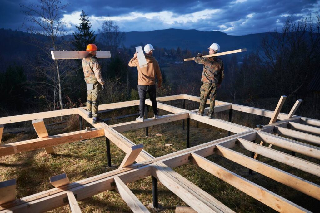 Men workers building wooden frame house on pile foundation. Back view of carpenters carrying wooden truss for timber framing. Carpentry concept.